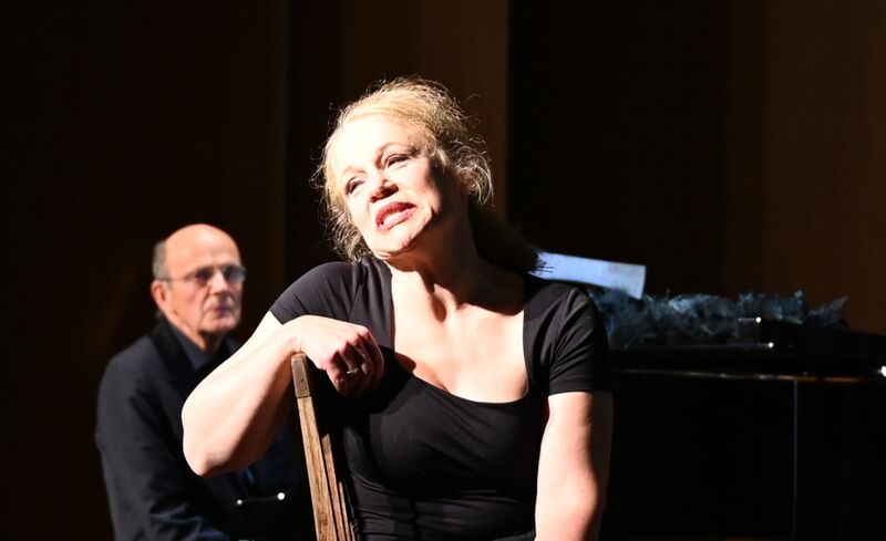 Die Sopranistin Kotzian sitzt auf einem Stuhl und spielt die Rolle der Ida Ehre, im Hintergrund der Pianist Christof Weber